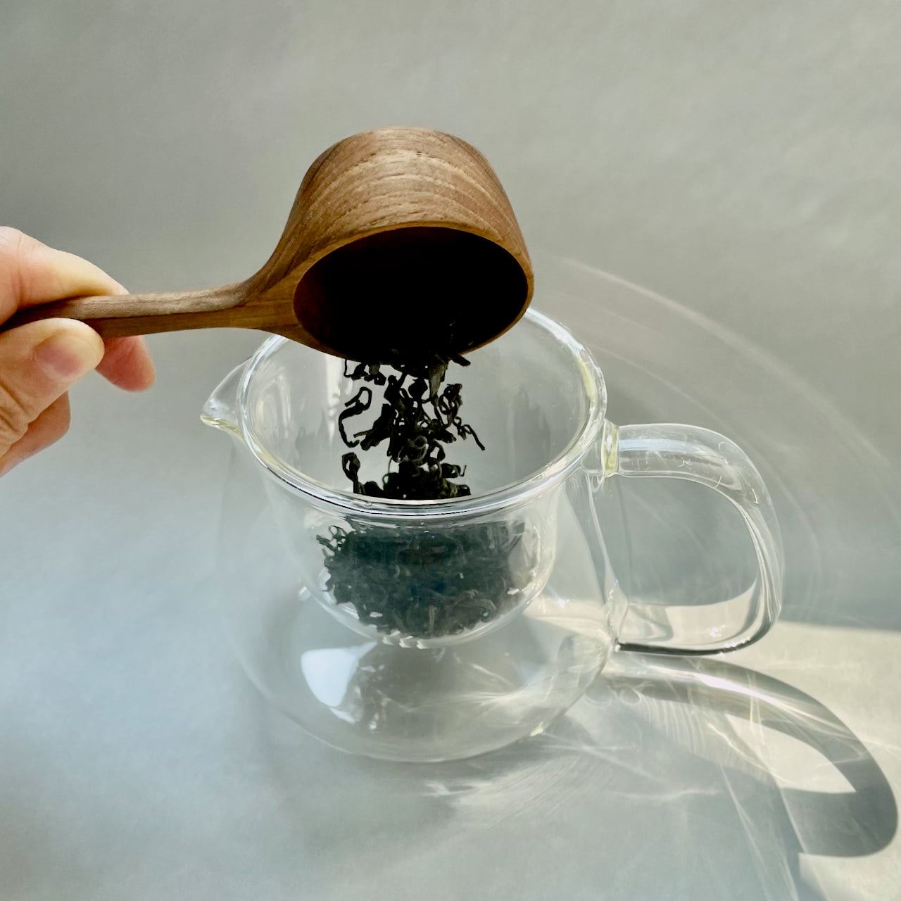 KINTO Tea Scoop offered by Wild rchid Tea, image of the wooden tea scoop with loose te leaves and glass tea kettle